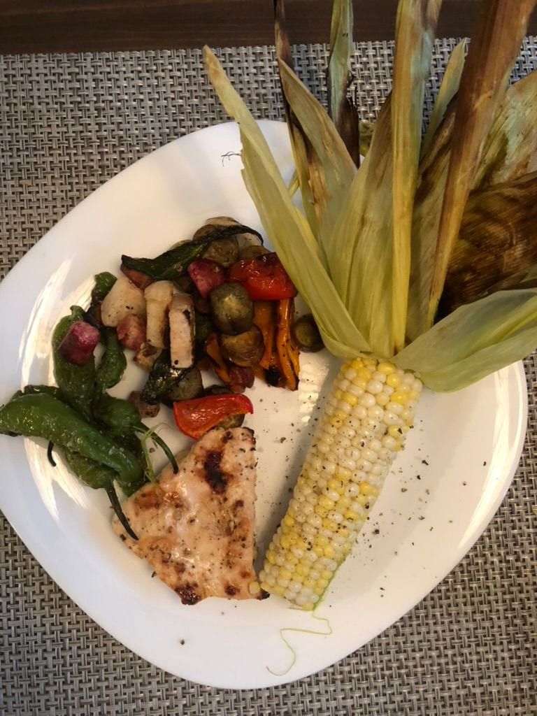 Grilled sweet corn in the husk on the plate, with blistered shishitos and roasted vegetables