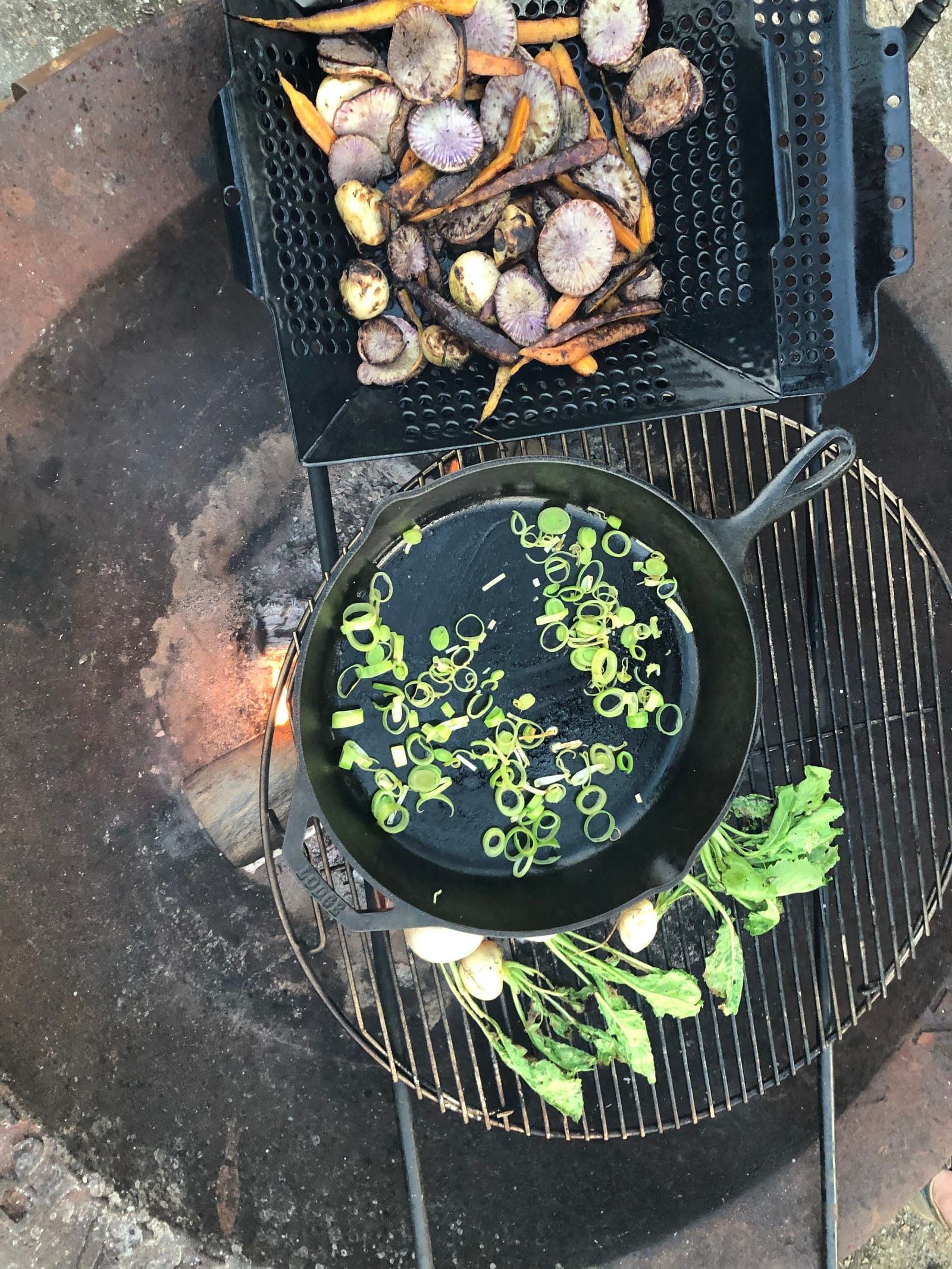 Turnips, carrots, leeks and radish going over open flame in a grill basket and a cast iron pan at the farm